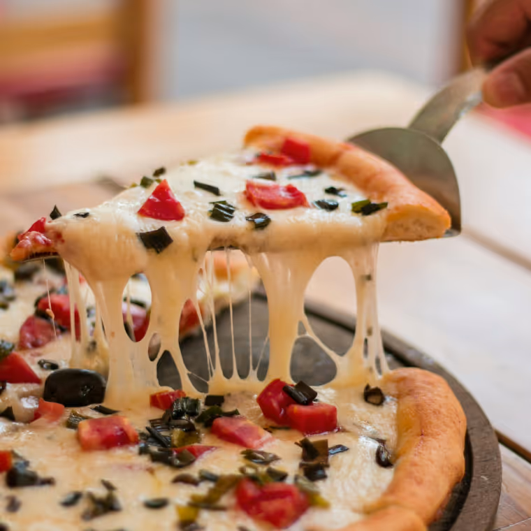 Slice of cheese pizza with tomatoes and herbs being lifted from a pan, showing melted, stretchy cheese.